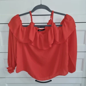 Korallröd volangblus från Gina Tricot - Trendig korallröd blus från Gina Tricot med volangdetaljer och axelband. Långärmad modell med lös passform och resår vid ärmslut. Perfekt för att addera färg till din outfit.