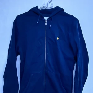 Mörkblå hoodie med dragkedja från Lyle & Scott - Mörkblå hoodie med dragkedja och huva från Lyle & Scott. Jackan har en gul broderad logga på bröstet och två fickor framtill. Perfekt för en avslappnad stil.