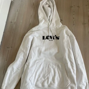 Vit hoodie från Levi's - Säljer en vit hoodie från Levi's med svart logga på bröstet. Tröjan har huva med dragsko och en stor magficka framtill. Passformen är relaxed och materialet känns mjukt och bekvämt. Perfekt för en avslappnad stil.