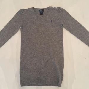 Grå stickad klänning från Polo Ralph Lauren - Säljer en grå stickad klänning från Polo Ralph Lauren med rund halsringning och tre dekorativa knappar på axeln. Tröjan har långa ärmar och en liten lila logga broderad på bröstet. Perfekt för lager-på-lager under kyliga dagar.