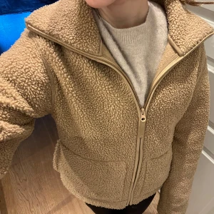 Brun teddyjacka med dragkedja - Mysig beige teddyjacka med hög krage och dragkedja framtill. Jackan har två stora fickor och är perfekt för kyliga dagar. Den har en avslappnad passform och är tillverkad i ett mjukt, fleece-liknande material. Inga skador.