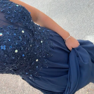 Balklänning med spets och öppen rygg - Säljer en marinblå långklänning med vackra spetsdetaljer och blommigt broderi upptill. Klänningen har öppen rygg och en elegant, svepande kjol. Perfekt för bal eller festliga tillfällen.