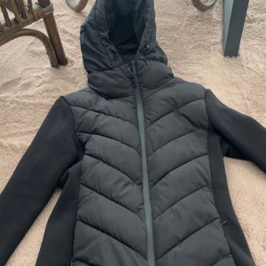 Svart pufferjacka med huva - Säljer en svart pufferjacka med huva och dragkedja. Jackan har quiltade partier framtill och baktill samt släta svarta ärmar. Perfekt för kyligare dagar och har en sportig look.