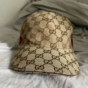 Beige keps - Säljer en beige keps i brunt över hela kepsen. På sidan finns ett grönt och rött band. Kepsen har böjd skärm och justerbar passform. Perfekt för dig som gillar lyxiga accessoarer.