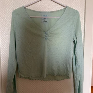 Ljusgrön v-ringad topp från Censored - Säljer en ljusgrön långärmad topp från Censored i storlek S. Toppen har v-ringning, små dekorativa knappar framtill och vågig kant nertill. Perfekt för dig som gillar en feminin och enkel stil.