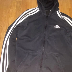 Svart Adidas jacka med huva och dragkedja - Svart Adidas jacka med vit logga på bröstet och klassiska vita ränder längs ärmarna. Jackan har huva, dragkedja framtill och två fickor. Perfekt för en sportig stil.