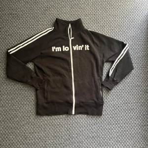 McDonald’s, om lovin’ it kofta - Svart track jacket med vit dragkedja och vita ränder längs ärmarna. Tryck med texten 'i'm lovin' it' på bröstet. Hög krage och två fickor med dragkedja. Tillverkad i bomullsmix, insida i mjuk fleece.
