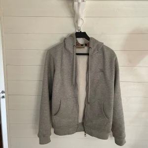 Grå hoodie från Burberry med klassiskt rutigt foder - Säljer en grå hoodie från Burberry med dragkedja och huva. Insidan har det ikoniska Burberry-rutiga fodret i beige, svart och rött. Två fickor framtill och ribbade muddar vid ärmslut och nederkant.