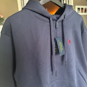 Mörkblå hoodie från Polo Ralph Lauren - Säljer en mörkblå hoodie från Polo Ralph Lauren med klassisk röd logga broderad på bröstet. Tröjan har huva med dragsko och en stor magficka framtill. Perfekt för en avslappnad och stilren look. Tveka inte på att höra av dig om du har några frågor!