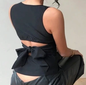 Svart ärmlös topp med rosett i ryggen - Säljer en svart ärmlös topp med snygg omlott och stor rosett i ryggen. Toppen har en tight passform och är perfekt för att skapa en trendig look. Materialet är mjukt och följsamt.