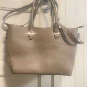 Beige axelväska med gulddetaljer - Stilren beige axelväska med både handtag och justerbar axelrem. Väskan har guldfärgade metalldetaljer och dragkedja upptill. Perfekt för att bära med sig det viktigaste till vardags. Enkel och klassisk design.