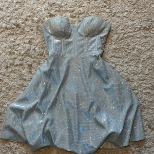 Glittrig axelbandslös silver/ljusblå klänning - Superfin axelbandslös kortklänning i glittrigt silvermaterial med markerad byst och lätt utställd kjol. Perfekt för dig som vill sticka ut på festen! Klänningen har en snygg passform och dragkedja bak.