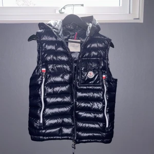 Navy blå dunväst från Moncler med huva - Snygg navy dunväst från Moncler med huva och två dragkedjefickor framtill. Västen har Moncler-logga på bröstet och vita detaljer vid dragkedjorna. Perfekt för lager-på-lager och stilren look.