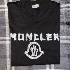Svart t-shirt från Moncler - Säljer en svart t-shirt från Moncler med stor vit logga och text framtill. Klassisk rund halsringning och mjukt material. Perfekt för dig som gillar streetwear och vill ha något stilrent men ändå med attityd.