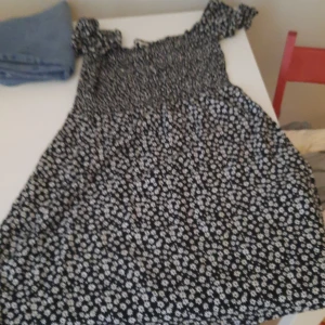 Svart blommig klänning med puffärm - Söt svart klänning med vitt blommönster och korta puffärmar. Klänningen har en smockad överdel och utställd kjol, perfekt för varma dagar. Lätt och luftig modell som passar till många tillfällen.