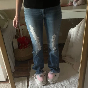 Blå slitna jeansbyxor - Säljer ett par blå jeansbyxor med slitningar framtill. Byxorna har normal passform och raka ben. Klassisk femficksmodell med knapp och dragkedja i gylfen. Perfekta för en avslappnad stil.