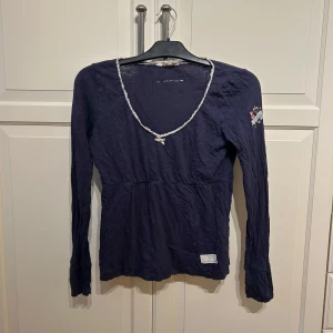 Mörkblå långärmad topp med spetskant från Odd Molly - Mörkblå långärmad topp från Odd Molly med v-ringning och dekorativ spetskant runt halsen. Liten broderad detalj på ena ärmen och en vit tygpatch nertill. Mjuk och tunn bomullskvalitet. I strl 1 (s)
