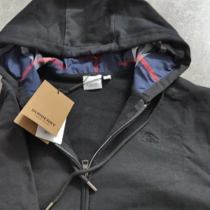 Svart Burberry Kofta - Säljer en svart hoodie från Burberry med dragkedja och justerbar huva. Insidan av huvan har Burberrys klassiska rutiga mönster i blått, rött och vitt. Broderad logga på bröstet och snygga metalldetaljer på dragkedjan.