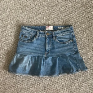 Jeanskjol med volang från Dobber - Säljer en blå jeanskjol från Dobber med volang nertill. Kjolen har klassiska jeansfickor fram och bak, knapp och dragkedja framtill samt detaljerade sömmar. 