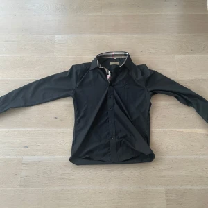 Svart skjorta från Burberry - Snygg svart skjorta från Burberry med klassisk krage och diskret broderad logga på bröstet. Skjortan har knäppning framtill och är långärmad. Perfekt för dig som gillar stilrena plagg med exklusiv känsla.