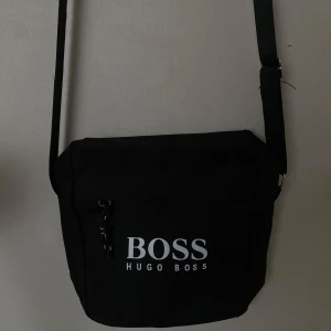 Svart axelväska från Hugo Boss - Säljer en svart axelväska från Hugo Boss med justerbar rem och tydlig logga framtill. Väskan har dragkedja och är perfekt för att bära med sig det viktigaste. Enkel och stilren design som passar till många tillfällen.