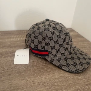 Navy/Beige keps från Gucci med monogram - Snygg navy/beige keps från Gucci med klassiskt GG-monogram i svart och röd-blå rand på sidan. Justerbar rem baktill i svart skinn. Perfekt accessoar för en trendig look.