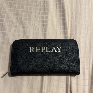 Svart plånbok från Replay - Säljer en svart plånbok från Replay med silverfärgad logga framtill. Plånboken har dragkedja runtom och flera fack inuti för kort och mynt. Het ny!