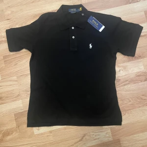 Polo Ralph Lauren pikétröja svart. - Klassisk svart pikétröja från Polo Ralph Lauren med vit broderad logga på bröstet. Tröjan har korta ärmar, krage och två knappar framtill. Perfekt för en stilren och tidlös look. Storlek S. Nyskick.