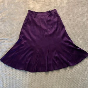 Lila satinkjol med dragkedja - Säljer en glansig lila kjol i satin med utsvängd form och dold dragkedja bak. Kjolen har bred linning och hällor för bälte. Perfekt för dig som vill ha en statement piece i garderoben!
