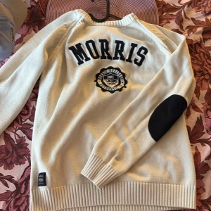 Vit stickad tröja från Morris - Vit stickad tröja från Morris med svarta bokstäver och emblem på ryggen. Tröjan har svarta förstärkningar på armbågarna och ribbade muddar vid ärmslut och nederkant. Perfekt för en stilren och klassisk look.