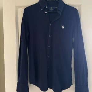Mörkblå långärmad skjorta från Ralph Lauren - Säljer en klassisk mörkblå skjorta från Ralph Lauren i modellen Featherweight Mesh. Skjortan har långa ärmar, knappar framtill och den ikoniska broderade loggan på bröstet. Perfekt för dig som gillar stilrena plagg med snygga detaljer.