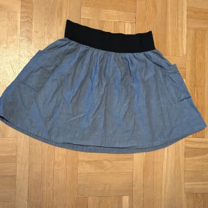 Blå kortkjol från JC med fickor - Supersöt blå kortkjol från JC med bred svart midjeresår och två stora fickor på sidorna. Kjolen har en avslappnad passform och är gjord i mjuk bomull, perfekt för en chill och stilren look.