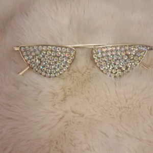 Glittriga kattögon-solglasögon - Unika solglasögon med kattögonform och massor av glittrande stenar på bågarna. Ramen är i guldton och stenarna skimrar i regnbågens färger. Perfekt accessoar för dig som vill sticka ut och glänsa extra mycket.