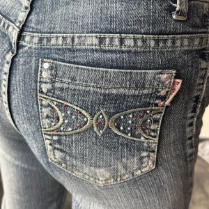 Bootcut/flare jeans med detaljer - Snygga blå bootcut jeans från en secoundhand butik med hög midja. Fickorna är dekorerade med broderier och strassdetaljer i flera färger som ger extra bling. Klassisk femficksmodell i jeansmaterial, perfekt för dig som vill sticka ut lite extra.