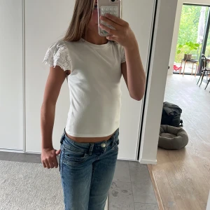 Vit topp med spetsärmar - Supersöt vit topp med korta ärmar i broderad spets och lätt ribbad struktur. Ärmarna har vågig kant och blommiga detaljer som ger en romantisk vibe. Perfekt till jeans eller kjol för en fräsch look.