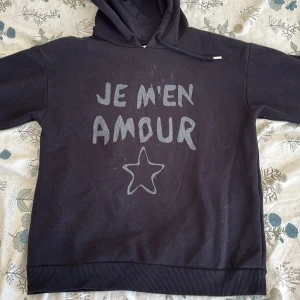 Svart hoodie Gina Tricot med tryck - Svart hoodie från Gina Tricot med texten 'JE M'EN AMOUR' och en stjärna i silvertryck på bröstet. Tröjan har huva, långa ärmar och ribbade muddar. Perfekt för dig som gillar en chill och trendig stil.