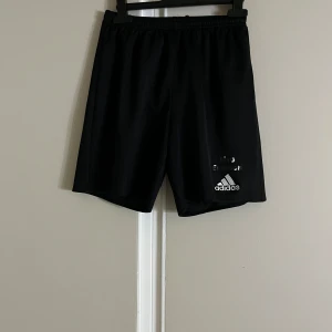 Svarta Adidas shorts climalite - Svarta sportiga shorts från Adidas med climalite-teknologi. Klassisk design med elastisk midja och Adidas-logga framtill. Perfekta för träning eller fotboll, tillverkade i lätt och snabbtorkande polyester.