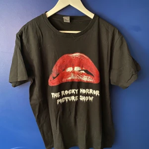 Rocky Horror svart t-shirt Gildan - Svart t-shirt från Gildan med tryck av röda läppar och texten 'The Rocky Horror Picture Show' på framsidan. Klassisk passform, rund hals och korta ärmar. Tillverkad i 100% bomull, perfekt för dig som gillar kultfilm och vill sticka ut.