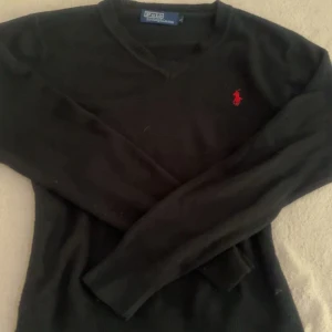 Svart stickad tröja Polo Ralph Lauren - Svart stickad tröja från Polo Ralph Lauren med klassisk v-ringning och röd broderad logga på bröstet. Långärmad modell i mjukt material som passar perfekt till jeans eller kjol. Enkel och stilren design som funkar året runt.