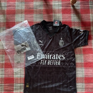 Svart AC Milan x Puma fotbollströja - Snygg svart AC Milan fotbollströja från Puma med diskret mönster och klassisk krage. Tröjan har korta ärmar, klubbmärke på bröstet och Emirates-sponsring framtill. Tillverkad i lätt och ventilerande material, perfekt för match eller träning.