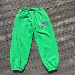 Gröna byxor från always do what you should do - Snygga gröna mjukisbyxor från always what you should do, elastisk midja och muddar vid benslut. Byxorna har ett vitt och svart tryck på ena benet och praktiska fickor, inklusive en bakficka med dragkedja. Perfekta för chill dagar eller träning.