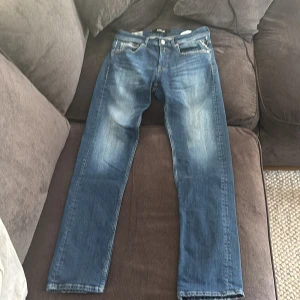 Replay Grover blå jeans straight fit - Snygga Replay Grover jeans i klassisk blå tvätt med raka ben och coola slitningar. Jeansen har fem fickor, kontrastsömmar och läderpatch bak i midjan. Perfekta för dig som gillar en tidlös och avslappnad look.
