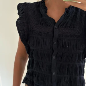 Marinblå blus med volang och knappar - Supersöt marinblå blus med volangdetaljer vid axlar och nederkant. Blusen har knappar framtill, lätt rynkad struktur och spetsdetaljer vid halsringningen. SLUTSÅLD på Nelly