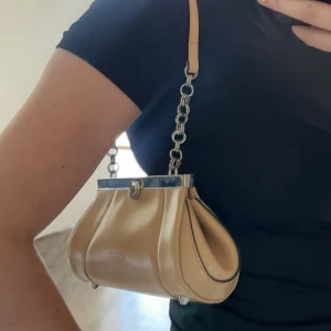 Beige axelväska från Estella i skinn - Snygg beige axelväska från Estella i glansigt skinn med cool silverkedja och metallram upptill. Väskan har unik form med markerade sömmar och små metallfötter undertill. Perfekt för dig som vill sticka ut med en trendig accessoar.