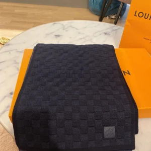 Mörkblå Louis Vuitton halsduk i ull - Snygg mörkblå halsduk från Louis Vuitton med diskret rutig struktur och liten läderpatch med logga. Tillverkad i 100% ull, perfekt för kalla dagar. Klassisk design som lyfter vilken outfit som helst.