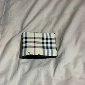 Rutig plånbok från Burberry - Snygg plånbok från Burberry med klassiskt rutigt mönster i beige, svart, vitt och rött. Utsidan har den ikoniska designen och insidan är i svart skinn med flera kortfack och plats för ID. Perfekt för dig som vill ha en stilren accessoar med lyxig känsla. Helt nytt skick!