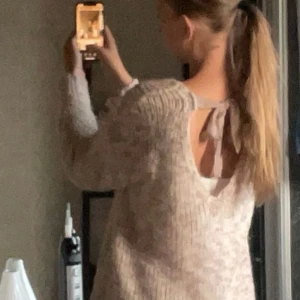 Beige stickad tröja med rosett i ryggen - Supermysig stickad tröja i beige och ljusa toner. Tröjan har lång ärm, grovstickat material och en snygg öppen rygg med rosett som detalj. Perfekt för lager-på-lager och chill dagar. Passar dig som gillar en lite loose fit och vill ha något både gulligt och stilrent.