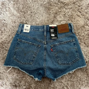 Levi's 501 jeansshorts - Säljer ett par helt oanvända klassiska Levi's 501 jeansshorts i blå denim. Alla lappar finns kvar! Modellen har råa, fransiga benslut och slitna detaljer framtill. Femficksmodell med knappgylf och bakfickor. Perfekta för sommaren!