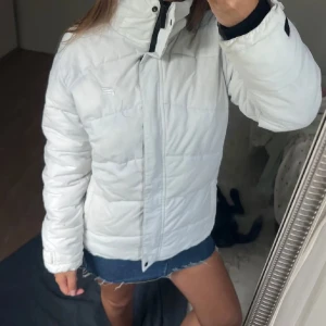 Vit dunjacka med hög krage - Snygg vit dunjacka med hög krage och diskret logga på bröstet. Jackan har quiltad design, dragkedja framtill och justerbara ärmslut. Perfekt för kalla dagar när du vill hålla stilen och värmen.