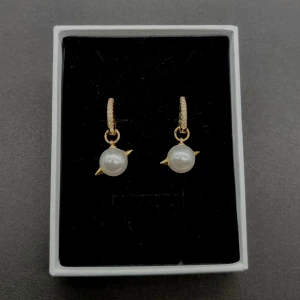 Guldörhängen med pärlor  - Snygga örhängen i guldton med gnistrande små stenar på ringen och hängande vita pärlor med coola gulddetaljer på sidorna. Perfekt för dig som vill ha något unikt och trendigt som sticker ut.